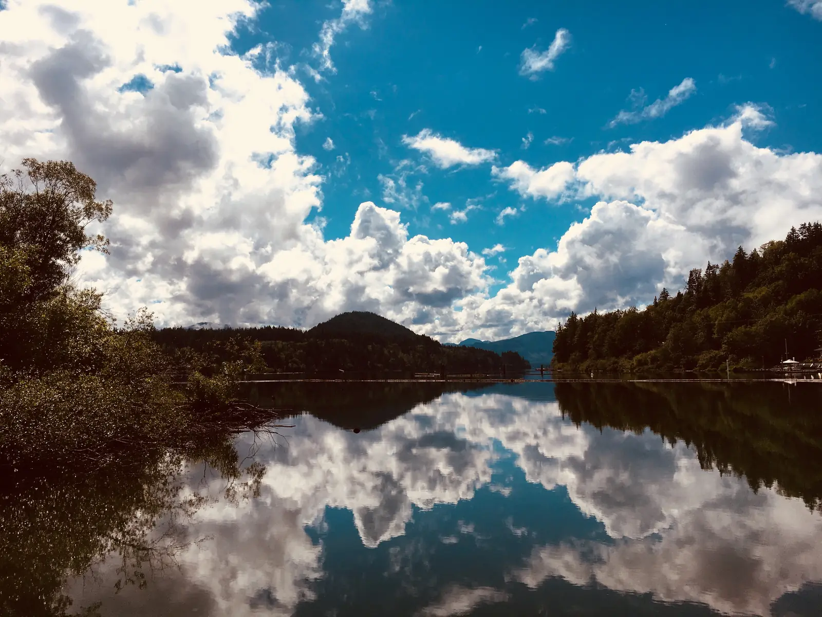 سحب وتلال مكسوّة بالغابات تنعكس على المياه الساكنة لبحيرة Cowichan Lake في بلدة Lake Cowichan